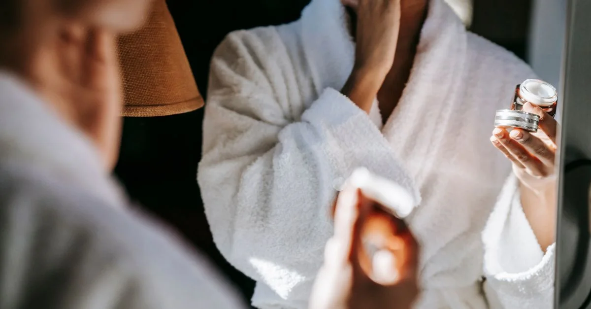 Femme non identifiable en peignoir appliquant de la crème hydratante devant un miroir.