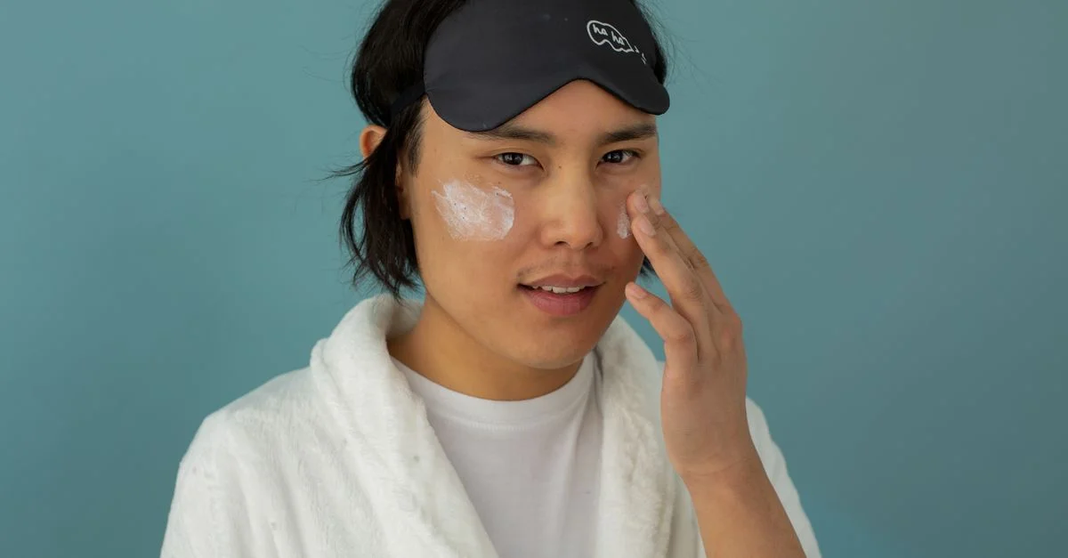 Homme asiatique en robe appliquant une crème de soin pour le visage avec un masque de sommeil.