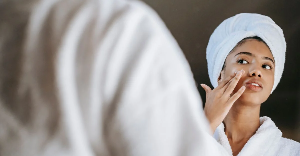 Une jeune femme applique de la crème pour la peau tout en portant un peignoir devant un miroir.