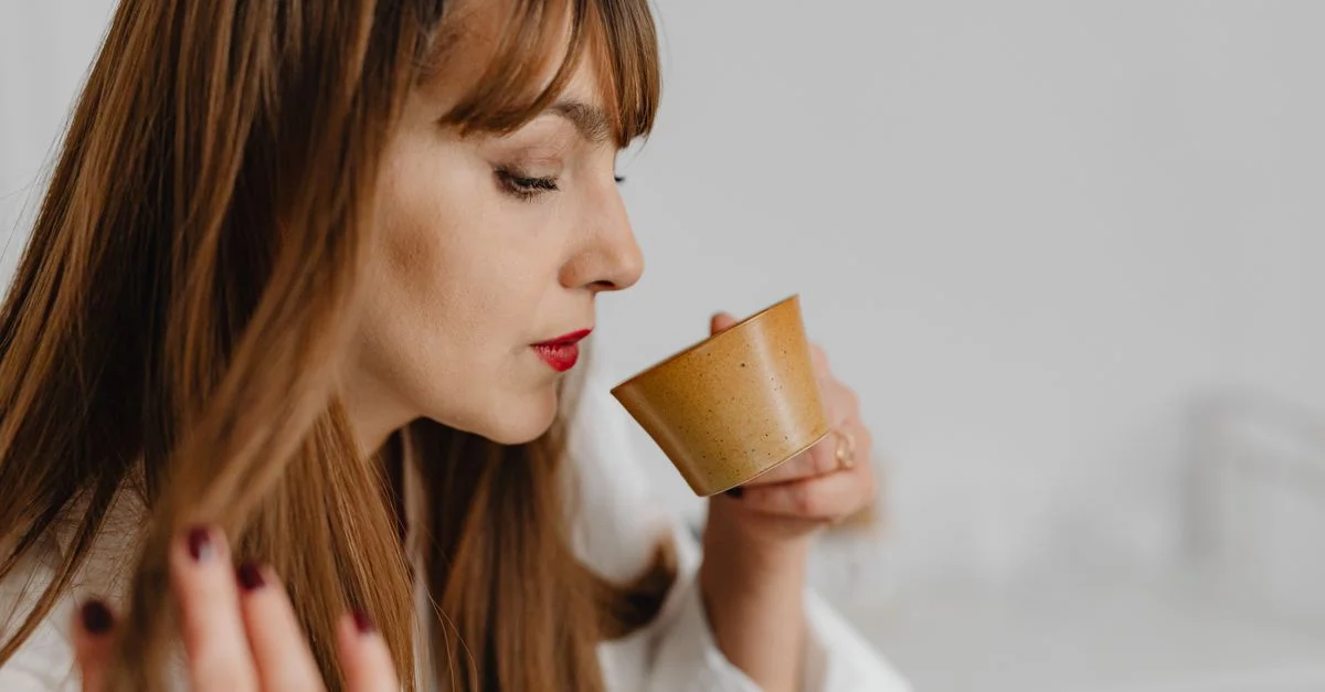 Une femme aux longs cheveux sirote du thé dans une tasse en céramique dans un cadre intérieur minimaliste.