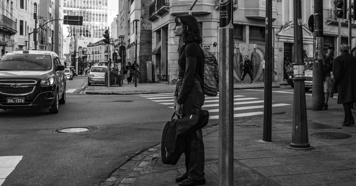 Scène de rue en noir et blanc avec une femme attendant à un passage piéton dans un cadre urbain.